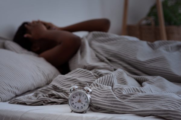 Sleepless, woman suffering from insomnia, sleep apnea or stress, headache or migraine. Tired and exhausted african american lady awake in middle of night, near alarm clock with time in dark, panorama