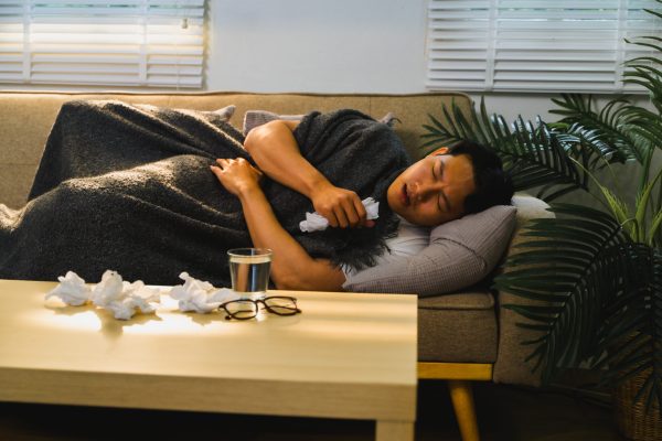 Asian man with a cold or flu is lying on the sofa at home, covered with a blanket, holding a tissue